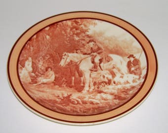 May include: A brown and white ceramic plate with a raised design of a group of people on horseback riding through a wooded area. The plate has a cream-colored background and a brown rim.