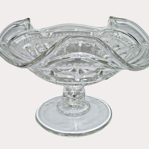 May include: Clear glass pedestal bowl with a ruffled rim. The bowl has a round base and a decorative stem. The rim has a scalloped edge. The bowl is likely used for serving desserts or displaying decorative items.