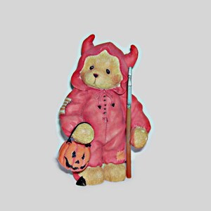 May include: A tan teddy bear figurine wearing a red hooded devil costume with horns. The bear holds a small orange jack-o'-lantern pumpkin bucket in one hand and a long, thin wooden stick with a silver tip in the other hand. The bear has black eyes.