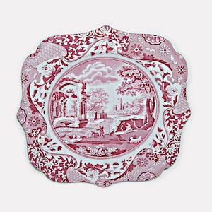 May include: A decorative, scalloped-edge plate with a red and white landscape design. The central image depicts a pastoral scene with figures, buildings, and trees. The border features floral and geometric patterns, creating a vintage aesthetic.