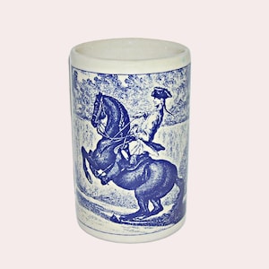 May include: A cylindrical ceramic mug with a blue and white toile design. The illustration depicts a person on horseback. The mug has a white rim and a light blue background.