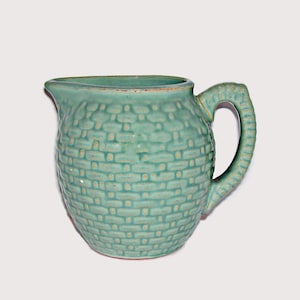May include: A light green ceramic pitcher featuring a textured basket-weave pattern across its exterior surface. The vessel includes a curved handle on the right side and a small pouring spout on the left, with a smooth rim and a glossy finish.