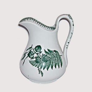 May include: A white ceramic pitcher featuring a dark green botanical transfer print of fern leaves and berry clusters on the front. The rim and handle are decorated with a matching dark green geometric pattern, creating a cohesive design.
