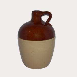 May include: A small, two-toned ceramic jug with a handle. The top portion is a glossy, dark brown, while the bottom is a matte, cream color. The jug has a rounded body and a small opening at the top.
