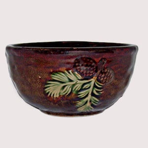 May include: A round ceramic bowl with a deep brown, glossy glaze. The exterior features a raised, textured relief design of a pine branch with two pinecones, colored in shades of green and brown, integrated into the surface of the bowl.