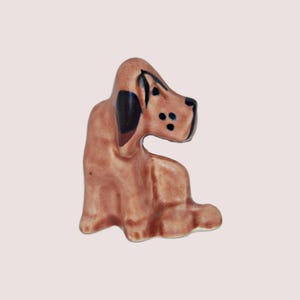 May include: A small ceramic figurine of a brown and black dog sitting with its head turned to the side. The dog has black ears and spots on its face.