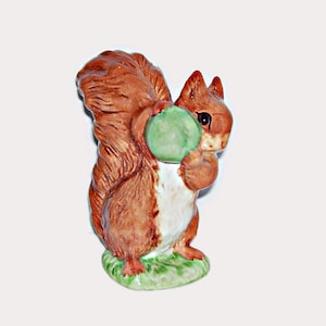 May include: A ceramic squirrel figurine featuring a brown coat with a white chest and a large, bushy tail. The squirrel is standing on a green base and is holding a round, light green object between its front paws. It has dark, glossy eyes.