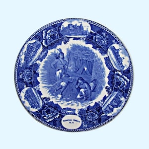 May include: Blue and white ceramic plate with a scene of Native Americans in a forest setting. The plate is decorated with a border of roses and buildings. The text "SARATOGA SPRINGS N.Y." is printed on the bottom of the plate.