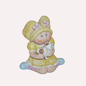 May include: A ceramic figurine of a young girl in a yellow dress and hat, holding a small white dog. The girl has blonde hair, blue eyes, and rosy cheeks. The figurine is detailed with pink accents and sits on a light blue base.