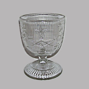 May include: A clear glass bowl with a footed base. The bowl has a decorative pattern of vertical lines and a floral design.