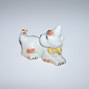 May include: A small white porcelain figurine of a dog with brown spots. The dog is lying down with its head turned to the side and its tail curled up. It is wearing a yellow bow.