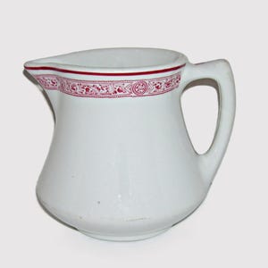 May include: White ceramic creamer with a red and white floral design around the rim.
