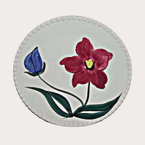 May include: A white ceramic plate with a hand-painted floral design. The plate features a large, open burgundy flower with a yellow center, a blue bud, and green leaves. The rim has a beaded edge.