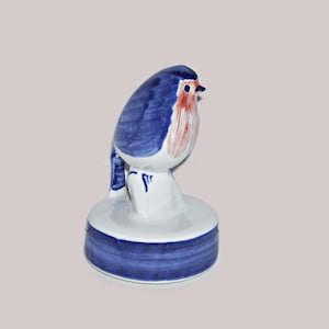 May include: A ceramic figurine of a bird perched on a circular base. The bird features a white body with blue painted wings and a reddish-orange chest. The circular base is white with a wide horizontal blue band painted around the lower edge.