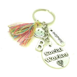 May include: Silver keychain with a colorful tassel, a smiley face charm, a letter B charm, and a charm that says "Social Worker" with a heart. The keychain also has a tag that says "Inspire".