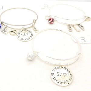 May include: Three silver charm bracelets with different charms. The first bracelet has a blue charm with white dots, a silver charm with a book, a silver charm with a lip, and a silver charm with the words "Everyone Deserves A Voice SLP". The second bracelet has a red charm with white dots and silver charms with the letters "SLP". The third bracelet has a silver charm with the words "Everyone Deserves A Voice SLP".