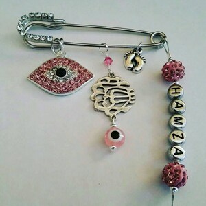 May include: A silver safety pin with pink and silver charms, including an evil eye, a charm with the word "Allah" in Arabic script, a charm with a footprint, and pink and silver beads that spell "HAMZA".