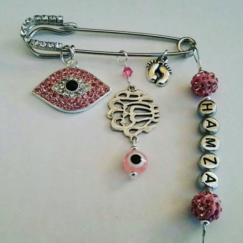 May include: A silver safety pin with pink and silver charms, including an evil eye, a charm with the word "Allah" in Arabic script, a charm with a footprint, and pink and silver beads that spell "HAMZA".