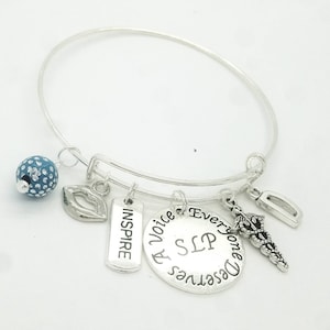 Könnte beinhalten: Silbernes Armband mit einem blauen Perlenanhänger, einem Lippenanhänger, einem Anhänger mit dem Wort "Inspire", einem Anhänger mit den Worten "Everyone Deserves A Voice", einem Anhänger mit den Buchstaben "SLP" und einem Caduceus-Anhänger.