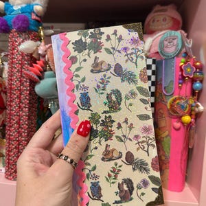 May include: A rectangular notebook featuring a cream-colored cover with a repeating pattern of rabbits, owls, squirrels, pinecones, and flowers. The cover has a holographic finish, a pink wavy trim along the left edge, and decorative metal corner guards.