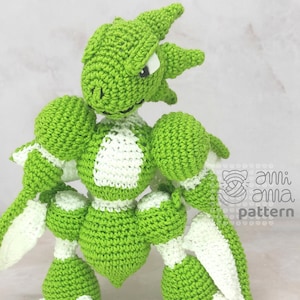 May include: Green and white crocheted amigurumi toy resembling a dragon with a spiky head and a segmented body. The toy has a white chest and white accents on its limbs.