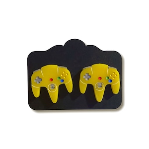 May include: A pair of yellow Nintendo 64 controller earrings. The controllers have gray buttons and are displayed on a black background.