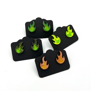Puede incluir: Pendientes de botón con forma de llama en tonos verdes, amarillos y naranjas. Cada par se exhibe en una tarjeta negra sobre un fondo blanco. Los pendientes tienen un acabado brillante y están diseñados para parecer llamas.