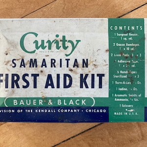 May include: A vintage metal first aid kit with a white background and blue and green accents. The kit is labeled "Curity Samaritan First Aid Kit" and "Bauer & Black, Division of the Kendall Company, Chicago". The contents of the kit are listed on the side, including surgical gauze, gauze bandages, adhesive tape, handi-tapes, burn-a-lay, iodine, aromatic spirits of ammonia, and scissors.