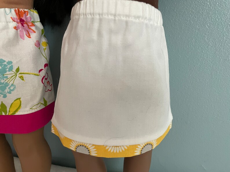 May include: Two white skirts with different colored trims. One skirt has a pink trim and the other has a yellow trim with a floral pattern.
