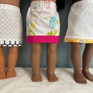 May include: Three dolls wearing different skirts. The doll on the left is wearing a white skirt with black polka dots and a black polka dot trim. The doll in the middle is wearing a white skirt with a floral print and a pink trim. The doll on the right is wearing a white skirt with a yellow floral print trim.