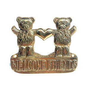 May include: A gold-toned metal wall plaque featuring two teddy bears standing side by side, holding a central heart between them. Below the bears, a rectangular base displays the embossed text "WELCOME FRIENDS" in capital letters.