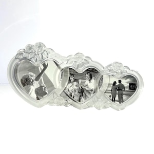 May include: A clear glass triple photo frame featuring three heart-shaped openings arranged horizontally. The frame is decorated with molded rose and leaf motifs at the top and bottom of each heart. The word Mikasa is printed on the right frame.