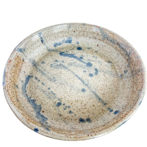 May include: A round ceramic bowl with a speckled beige and tan base. The interior features a glossy finish with irregular blue paint splatters and streaks that extend from the center toward the raised, slightly textured rim of the dish.