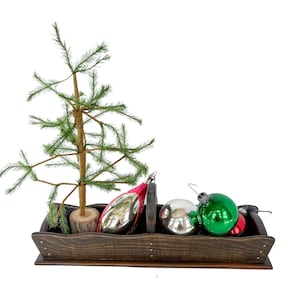May include: A dark brown wooden rectangular tray with a central handle, containing a small artificial pine tree on a wood slice base, a silver teardrop ornament, a silver spherical ornament, and a shiny green spherical ornament.