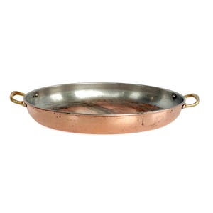 May include: A shallow, round copper pan with a metallic silver-toned interior lining. The exterior features a warm, polished copper finish with visible signs of use. Two small, curved brass handles are attached to opposite sides of the rim.