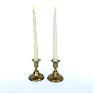 May include: Two matching brass candlesticks with a polished metallic finish, each featuring a wide circular base and a slender stem. Each candlestick holds a tall, thin, off-white taper candle with a small, glowing yellow flame at the top.