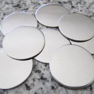 May include: A group of eight silver-colored metal discs, stacked and overlapping on a gray speckled surface.