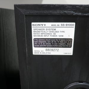 May include: A black Sony SS-B1000 speaker system with a wood grain texture. A white rectangular label on the back displays technical specifications including 8 ohm impedance, 120W maximum input power, serial number 8809272, and Made in Malaysia.