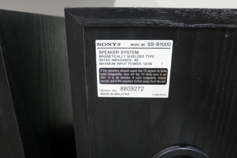 May include: A black Sony SS-B1000 speaker system with a wood grain texture. A white rectangular label on the back displays technical specifications including 8 ohm impedance, 120W maximum input power, serial number 8809272, and Made in Malaysia.