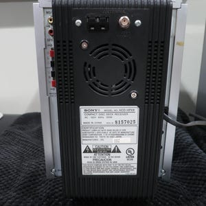 以下が含まれることがあります： シルバーとブラックのソニー製HCD-HPX9コンパクトディスクデッキレシーバーの背面パネル。中央には黒いプラスチック製の冷却ファン用グリル、アンテナポート、技術仕様とシリアルナンバーが記載された白い識別ラベルが配置されています。