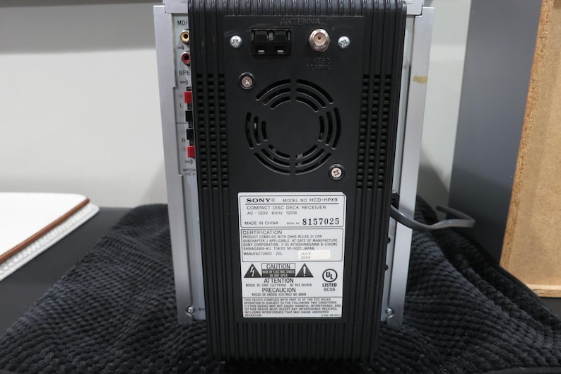 以下が含まれることがあります： シルバーとブラックのソニー製HCD-HPX9コンパクトディスクデッキレシーバーの背面パネル。中央には黒いプラスチック製の冷却ファン用グリル、アンテナポート、技術仕様とシリアルナンバーが記載された白い識別ラベルが配置されています。