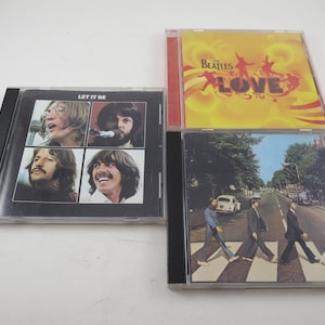May include: Four Beatles CD albums. The "Let It Be" album cover features four individual portraits of the band members. The "Love" album has a yellow cover with silhouettes. The "Abbey Road" album shows the band crossing a street.