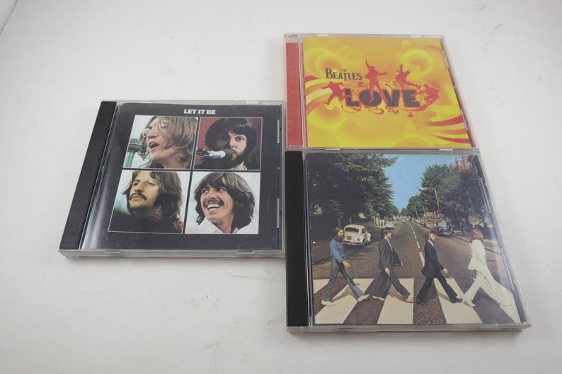 May include: Four Beatles CD albums. The "Let It Be" album cover features four individual portraits of the band members. The "Love" album has a yellow cover with silhouettes. The "Abbey Road" album shows the band crossing a street.