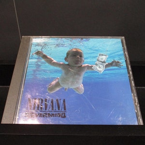 May include: A CD case with a blue and white image of a baby swimming underwater holding a dollar bill. The text "Nirvana" and "Nevermind" is printed on the case.