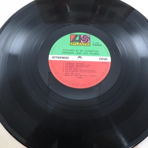 Puede incluir: Un disco de vinilo negro con una etiqueta roja y verde. La etiqueta dice "Pictures at an Exhibition" de Emerson, Lake and Palmer. La grabaci&oacute;n es est&eacute;reo y est&aacute; etiquetada como "One".