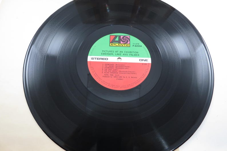 Puede incluir: Un disco de vinilo negro con una etiqueta roja y verde. La etiqueta dice "Pictures at an Exhibition" de Emerson, Lake and Palmer. La grabaci&oacute;n es est&eacute;reo y est&aacute; etiquetada como "One".