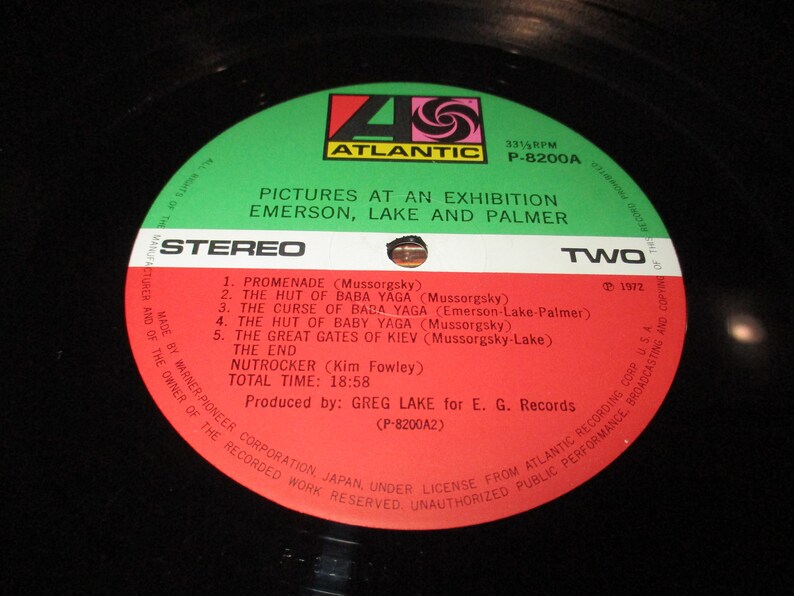 Op de afbeelding: Een rood en wit vinyl platenlabel met de tekst "Pictures at an Exhibition Emerson, Lake and Palmer" en "Stereo" in zwart. Het label bevat ook de tekst "Two" en "1972" in wit. Het platenlabel is groen aan de buitenrand.