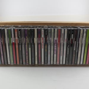May include: A wooden CD storage box filled with a collection of music CDs. The CDs are in their jewel cases, with various album covers visible. The box is made of light-colored wood.