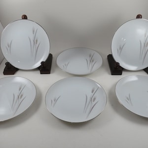 May include: A set of five white ceramic plates featuring a delicate gray wheat stalk pattern and a thin silver rim. Two of the plates are displayed upright on dark wooden stands, while the remaining three plates are arranged flat in front.