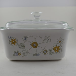 May include: White rectangular casserole dish with a clear glass lid. The dish is decorated with a floral pattern in yellow, blue, and green. The lid has a clear glass knob. A vintage kitchenware item.